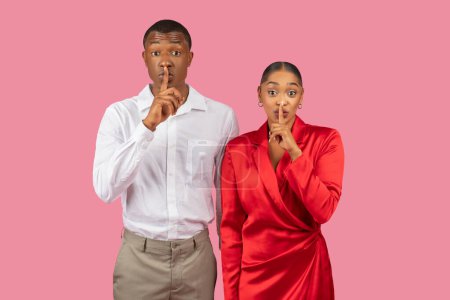 Photo for Black man and woman in elegant attires with fingers on their lips, giving hushing gesture, implying need for silence or secrecy, against pink backdrop - Royalty Free Image