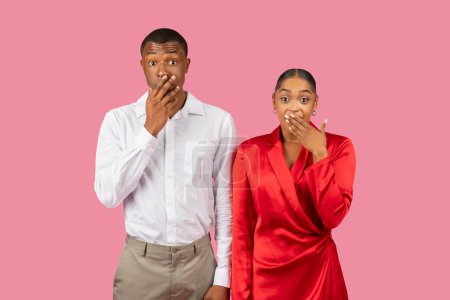 Astonished black couple covering mouths, man in white shirt and woman in red dress express surprise, set against pink background