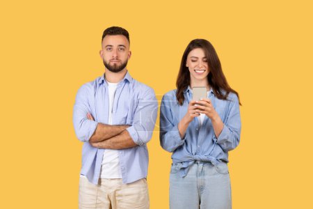 Photo for Skeptical man stands with arms crossed beside smiling woman engrossed in texting on her smartphone, both in blue shirts, on yellow backdrop - Royalty Free Image