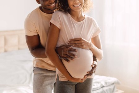 Téléchargez les photos : Concept de grossesse. Coup de feu recadré de jeune couple afro-américain en attente de bébé, mari embrassant sa femme enceinte et touchant le ventre en pleine croissance dans la chambre à coucher à l'intérieur - en image libre de droit