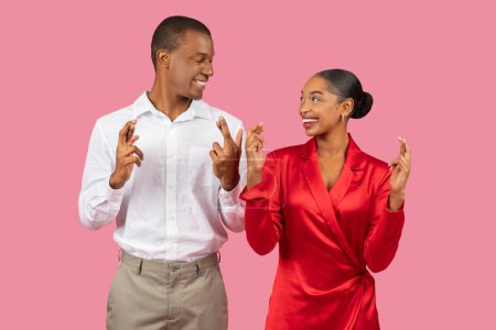 Foto de Hombre negro alegre en camisa y mujer encantada en vestido que cruza sus dedos para la buena suerte, compartiendo mirada esperanzadora contra el fondo rosa - Imagen libre de derechos