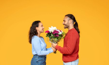 Foto de Una mujer radiante en un top azul sonríe agradecida mientras recibe un ramo de flores vívidas de un hombre igualmente feliz en un suéter rojo, sobre un fondo amarillo brillante. - Imagen libre de derechos