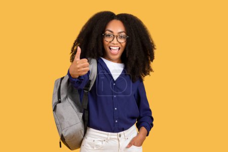 Photo for Cheerful black lady student wearing glasses and backpack gestures thumbs up, set against yellow background, embodying positivity and approval in an academic setting - Royalty Free Image