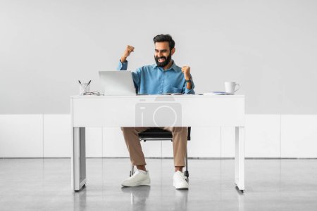 Foto de Hombre profesional indio eufórico con los brazos levantados que muestra el júbilo en el éxito del trabajo, sentado mientras que el escritorio moderno con el ordenador portátil y los documentos, longitud completa - Imagen libre de derechos