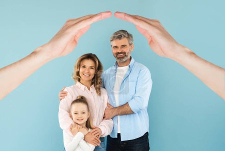 Photo for Middle aged parents posing with their cute daughter under hands made roof above their heads, blue studio background, collage. Love, parenthood, affection concept - Royalty Free Image