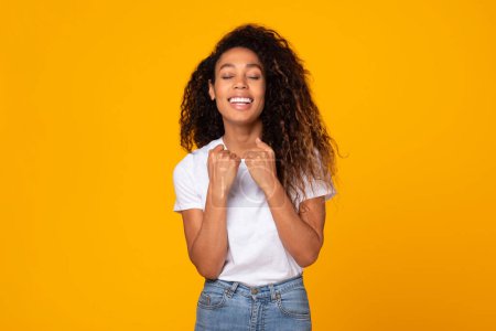 Téléchargez les photos : Curly jeune femme afro-américaine geste oui dans la célébration du succès et de la victoire, posant les yeux fermés en studio sur fond jaune. Gagnant, concept de réalisation - en image libre de droit