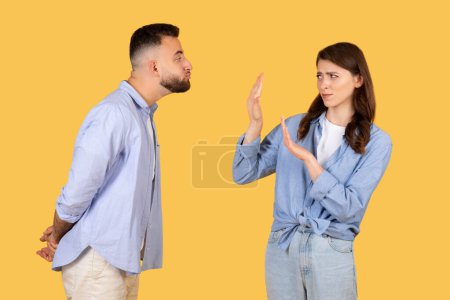 Man trying to kiss while woman firmly displays stop hand sign, indicating rejection or boundary setting, against vibrant yellow backdrop