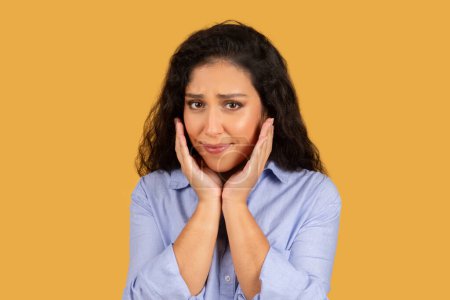 Photo for Worried arab young woman with curly hair holding her face in hands, looking at the camera with a troubled expression on a simple yellow studio background, close up - Royalty Free Image