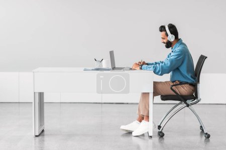 Téléchargez les photos : Homme barbu indien professionnel profondément absorbé dans son travail, en utilisant des écouteurs à élégant, bureau minimaliste avec ordinateur portable, incarnant flux de travail productif, vue latérale - en image libre de droit