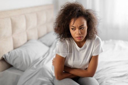 Téléchargez les photos : Jeune femme afro-américaine déprimée se câlinant souffrant de dépression et se sentant mal, ayant un problème d'estomac ou des périodes douloureuses, assis dans la chambre moderne à la maison - en image libre de droit