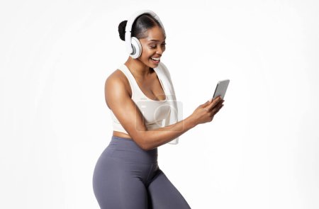 Smiling black woman in sportswear with headphones listens song on phone, isolated on white background, studio shot. Workout motivation and favorite music for fitness training