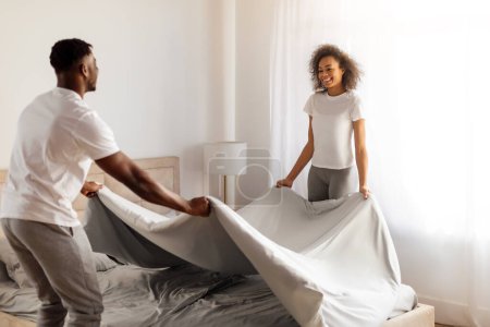 Téléchargez les photos : Jeune couple afro-américain couvrant lit avec couverture ensemble, rangeant leur chambre le matin, commençant la journée avec nettoyage de routine. Concept de confort domestique - en image libre de droit