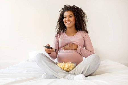 Foto de Feliz señora embarazada negra relajándose en casa, viendo la televisión y comiendo palomitas de maíz en la cama, sonriente mujer afroamericana embarazada sosteniendo el mando a distancia, Descanso en el acogedor dormitorio, Copiar el espacio - Imagen libre de derechos