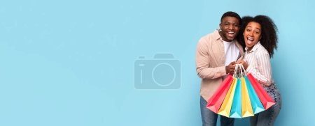 Téléchargez les photos : Ravi couple afro-américain serrant sacs à provisions colorés, exprimant le bonheur et le plaisir des consommateurs sur fond bleu calme, panorama avec espace de copie - en image libre de droit