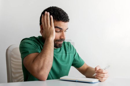 Téléchargez les photos : Homme frustré avec une barbe dans un t-shirt vert tenant sa tête d'une main tout en écrivant dans un carnet, assis à un bureau blanc, montrant des signes de stress ou de concentration - en image libre de droit