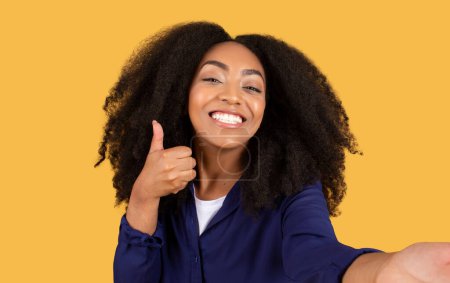 Téléchargez les photos : Radieuse jeune femme noire aux cheveux bouclés sourit largement et montre les pouces vers le haut geste, posant pour selfie sur une toile de fond jaune vif - en image libre de droit