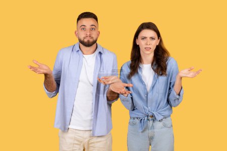 Photo for Perplexed young man and woman shrugging with palms up, displaying expressions of doubt and confusion, standing together against plain yellow background - Royalty Free Image