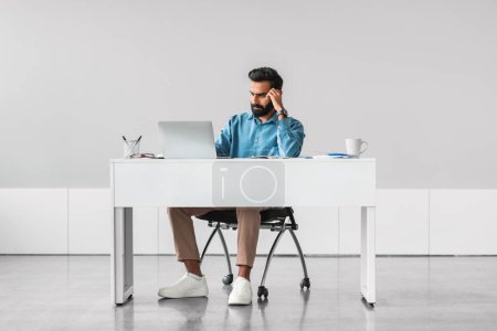Photo for Pensive bearded indian businessman in blue shirt, deep in thought at his minimalist workstation with laptop, in bright and modern office space - Royalty Free Image