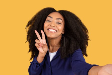 Téléchargez les photos : Joyeuse dame noire aux cheveux bouclés, prend selfie et clignote geste de paix, incarnant l'esprit de la jeunesse sur fond jaune vif, capturant un moment de vie étudiante insouciante - en image libre de droit