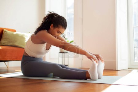 Téléchargez les photos : Dame sportive assise sur un tapis de yoga pratiquant des pilates effectuant des exercices d'aérobic tactile sur les orteils, montrant sa force physique et sa flexibilité dans le salon à l'intérieur. - en image libre de droit