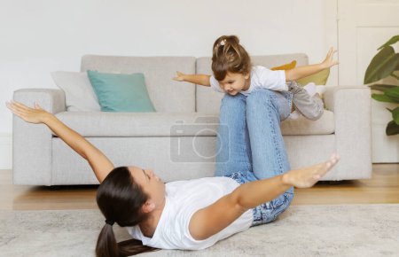 Photo for Mom and little daughter having fun together at home, mother lying on her back with arms extended, lifting her delighted child in an airplane game, playing and laughing in living room interior - Royalty Free Image