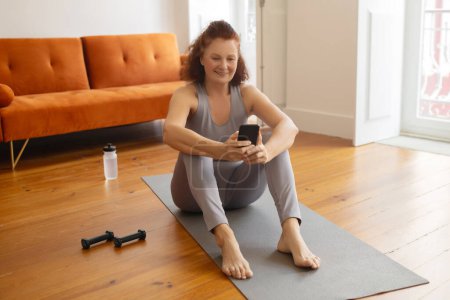 Photo for Smiling senior woman sitting on yoga mat and using her smartphone, happy elderly female taking break after fitness training at home, enjoying sport tracker application on mobile phone, copy space - Royalty Free Image