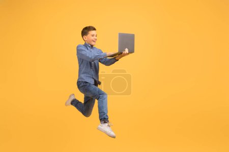 Téléchargez les photos : Adolescent excité garçon avec ordinateur portable dans les mains sautant sur fond jaune, en mouvement coup de jeune homme heureux regardant l'écran de l'appareil avec intérêt, bénéficiant d'une offre en ligne, pleine longueur - en image libre de droit