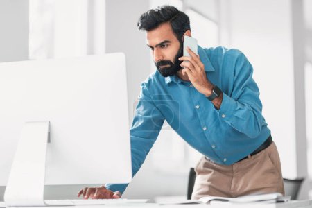 Konzentrierter indischer Geschäftsmann in blauem Hemd auf Abruf, während er gleichzeitig am Computer arbeitet und effektives Multitasking im Büro zeigt