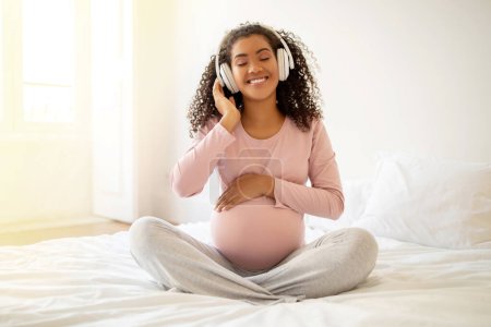 Téléchargez les photos : Heureuse femme enceinte noire assise sur le lit, écoutant de la musique et touchant le ventre, souriante femme afro-américaine dans des écouteurs sans fil se détendre dans la chambre, profiter du temps de grossesse, copier l'espace - en image libre de droit