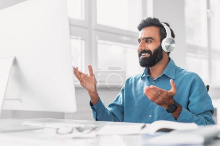 Photo for Engaged indian bearded man in blue shirt with headphones gesticulating, fully immersed in lively virtual conversation in bright office setting - Royalty Free Image