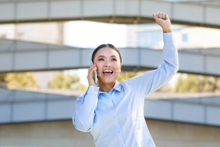 Japanese entrepreneur woman talking on her phone triumphantly shaking fist, standing in front of business center building outdoors, symbolizing career victory in modern city
