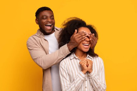 Foto de Feliz pareja afroamericana en ropa casual, hombre cubriendo los ojos de las mujeres para una alegre sorpresa sobre un vibrante fondo amarillo, ambos riendo con emoción - Imagen libre de derechos