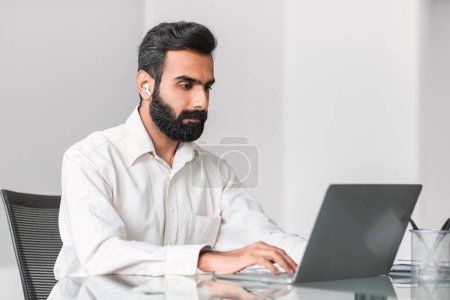 Professioneller indischer Mann in knackigem weißem Hemd, der Kopfhörer benutzt, während er am gläsernen Schreibtisch intensiv am Laptop arbeitet und die moderne Büroproduktivität verkörpert