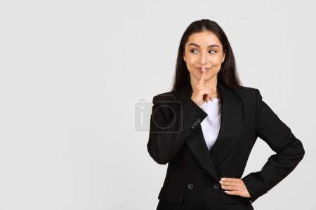 Photo for Confident businesswoman in a sleek black suit places her finger on her lips in a gesture for silence, standing against neutral grey background, free space - Royalty Free Image