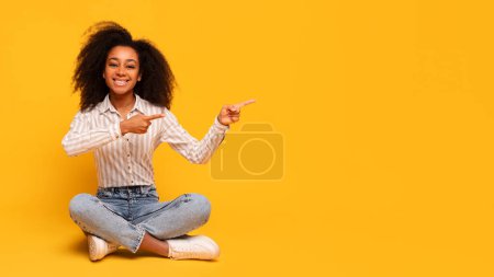 Téléchargez les photos : Jeune femme afro-américaine excitée avec les cheveux bouclés assis jambes croisées, pointant vers la droite à l'espace de copie, sur fond jaune vif, panorama - en image libre de droit