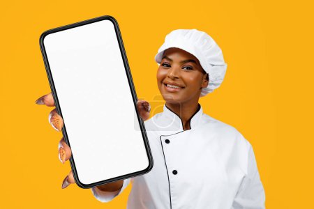 Téléchargez les photos : Belle femme noire chef posant sur fond jaune, joyeuse cuisinière afro-américaine dame montrant Big Phone avec écran blanc, Mockup, Copier l'espace. Blog culinaire alimentaire en ligne - en image libre de droit