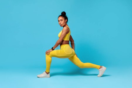Photo for Workout Training. African American fitness lady stretching legs lunging forward, wearing yellow fitwear over blue studio background, embodying strength and wellness. - Royalty Free Image