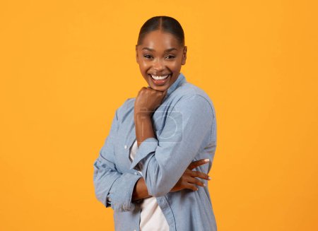 Photo for Black millennial lady standing touching chin with cheerful smile, looking at camera with confident expression against yellow studio backdrop. Female beauty and confidence - Royalty Free Image