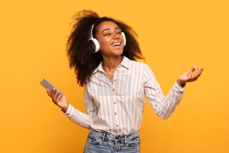 Téléchargez les photos : Jeune femme noire aux cheveux bouclés sentant la musique, portant un casque et tenant un smartphone, les yeux fermés et la main levée en geste paisible sur fond jaune - en image libre de droit