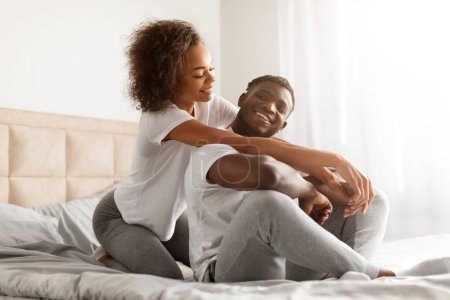 Romantischer Morgen. Lächelndes schwarzes junges Paar genießt Romantik und Intimität im modernen Schlafzimmer, liebende Frau umarmt ihren Mann, der drinnen auf dem Bett sitzt. Glückliche Beziehung, Ehe