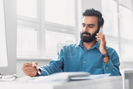 Téléchargez les photos : Homme d'affaires indien attentif en chemise bleue lors d'un appel téléphonique sérieux, stylo à la main, examinant attentivement les documents dans son espace de travail bien organisé - en image libre de droit