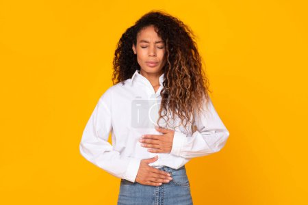 Photo for Gastritis Disease. Black young woman touching stomach while having painful spasm in belly, suffering from stomachache, standing over yellow studio background. Abdominal pain symptom, health problem - Royalty Free Image