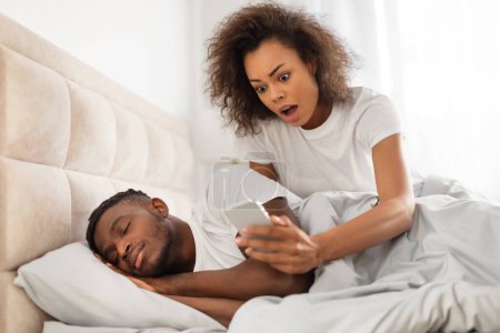 Infidelity, jealousy issue. Distrustful black woman checks her boyfriends cellphone and reading messages suspecting cheating, peeking while man peacefully sleeping in bed in modern bedroom at home