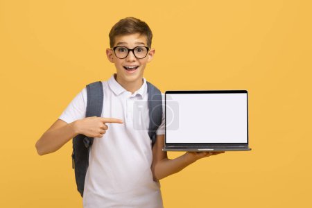 Photo for Enthusiastic schoolboy wearing glasses pointing at blank white laptop screen, happy teen male kid with backpack standing on yellow background, symbolizing digital learning or online education, mockup - Royalty Free Image
