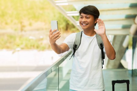 Photo for Positive chinese guy tourist have video call while waiting for plane at airport, using smartphone, waving and smiling at phone screen. Travel blogger streaming from train station. Tourism, traveling - Royalty Free Image