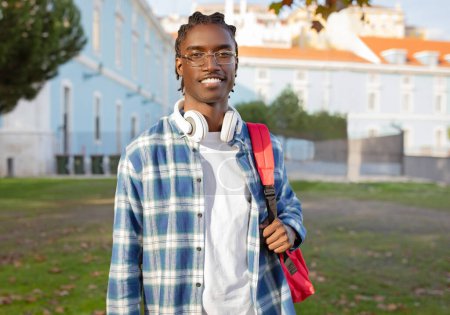 Téléchargez les photos : Souriant étudiant afro-américain confiant avec casque autour du cou et sac à dos rouge vif debout à l'extérieur campus. Diplôme universitaire, concept d'éducation - en image libre de droit