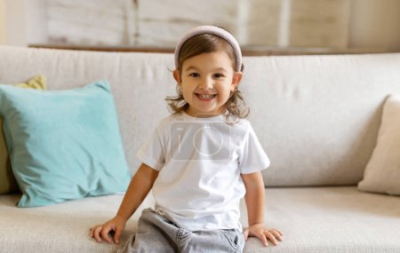 Photo for Happy preschool little girl comfortably seated on couch at home, cute female child smiling at camera, relaxing on comfortable sofa, expressing carefree childhood in homely setting, copy space - Royalty Free Image