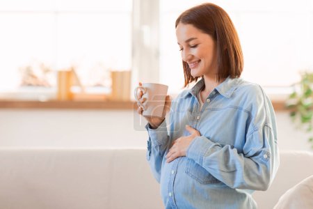 Téléchargez les photos : Jeune femme enceinte appréciant boire du thé chaud et toucher le ventre debout à l'intérieur, tenant la tasse. Femme enceinte ayant une boisson saine ou un café le matin. Espace libre - en image libre de droit