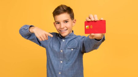 Great Bank. Happy Teen Boy Pointing At Credit Card In Hand And Looking At Camera, Cheerful Smiling Male Teenager Advertising Easy Payments, Standing Over Yellow Studio Background, Copy Space