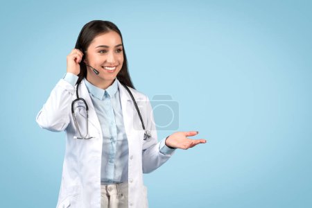 Smiling young female doctor wearing headset, making welcoming gesture, dressed in white coat, prepared for teleconsultation on blue background, free space